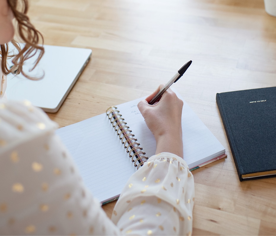 Over the shoulder shot of a person wearing a cream and gold blouse writing in a small notebook on a wood block table.