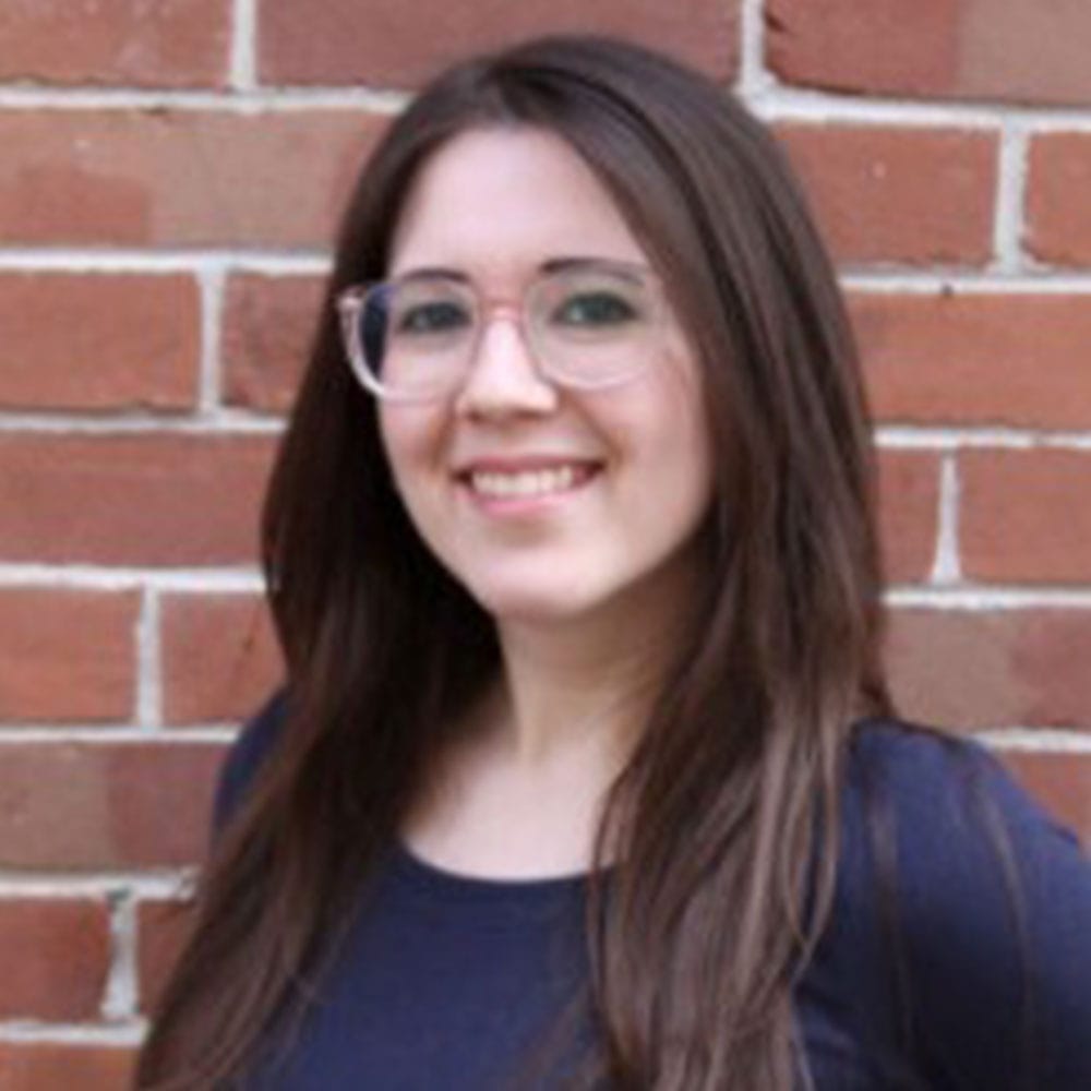 Jessica, our editor-in-chief, wears rose-colored plastic glasses and a navy shirt. She has long brown hair and is pictured smiling in front of a red brick wall.
