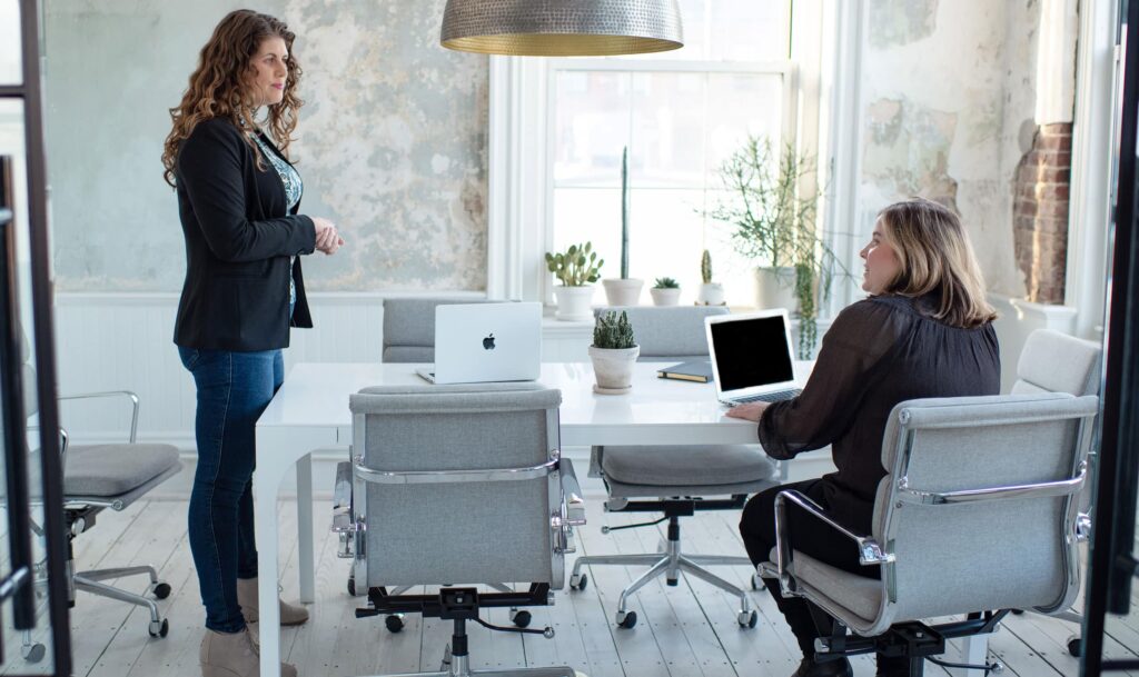 A woman stands in a chicly-decorated conference room at a modern content marketing agency, speaking to a colleague seated in a gray tweed chair at the opposite end of the white conference table.