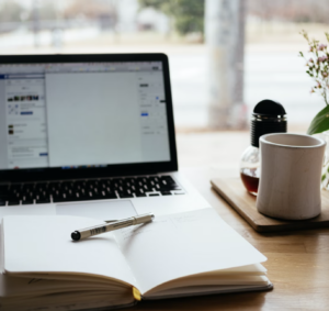 Notebook and pen open in front of a laptop next to a cup of coffee