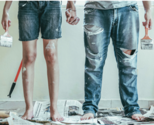 couple holding hands and holding paintbrushes over dropcloth with paint splattered on their legs